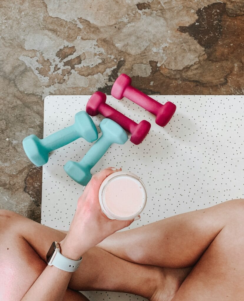 Woman sitting on a yoga mat with dumbbells and drinking a protein shake after fitness training